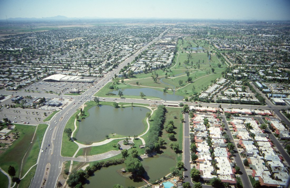 Aerial view of a cityscape with a large park featuring multiple lakes, surrounded by residential neighborhoods and commercial areas.