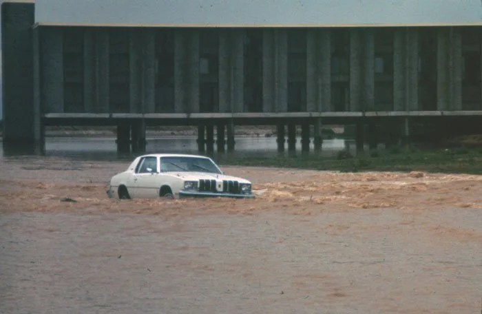 A white car partially submerged in floodwaters with muddy water surrounding it.