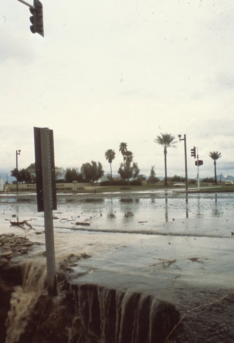 Flooded street with waterfall near a traffic signal, with palm trees and cloudy sky in the background.