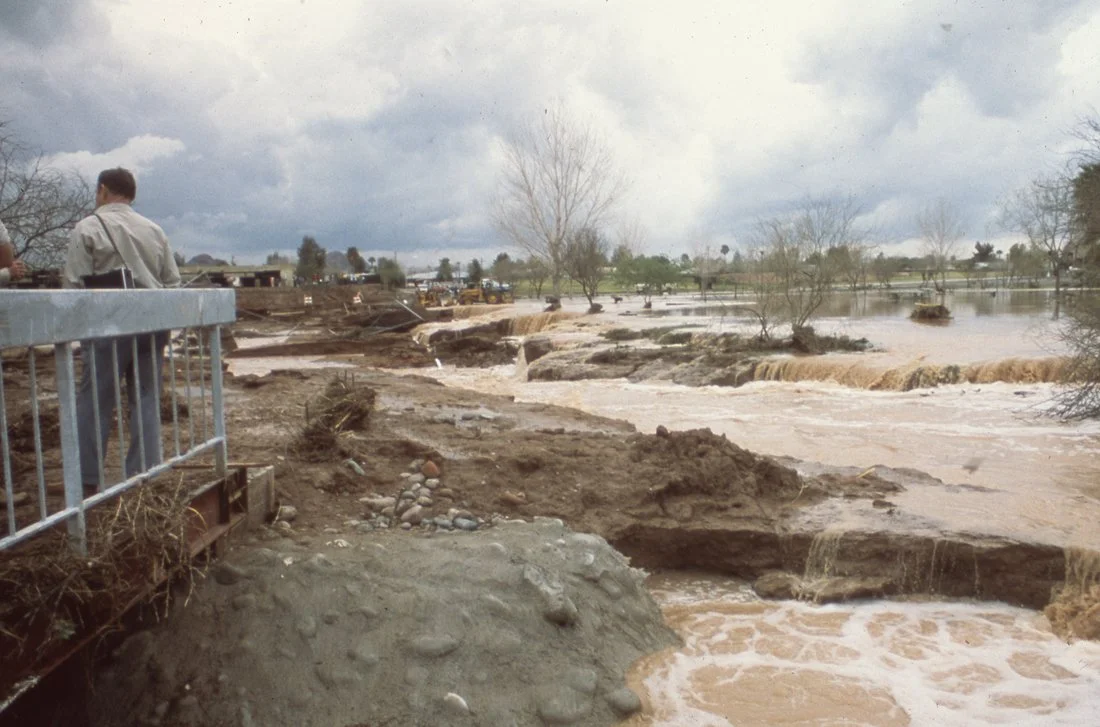 Floodwaters rushing over a damaged riverbank with muddy water and eroded land, cloudy sky, and trees in the background.