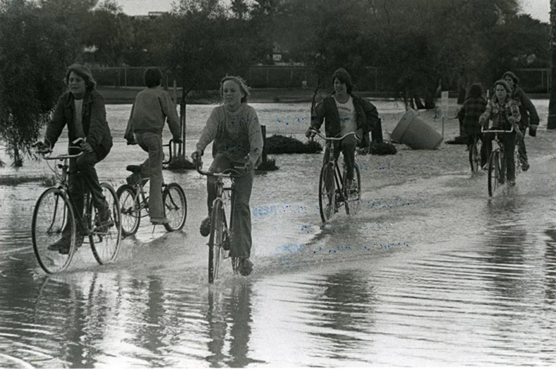 A group of children riding bicycles through a flooded area with water at ankle level, some crossing the water and some on the sidewalk nearby.