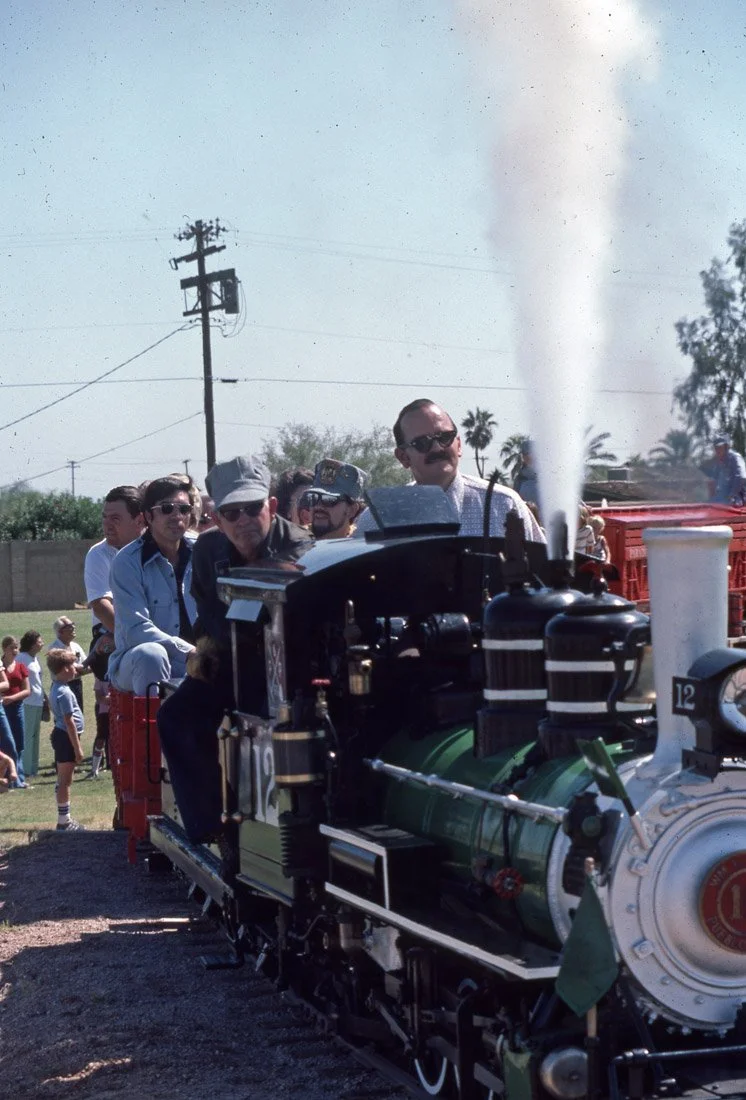 People riding on a miniature train during a sunny outdoor event with onlookers and children in the background.