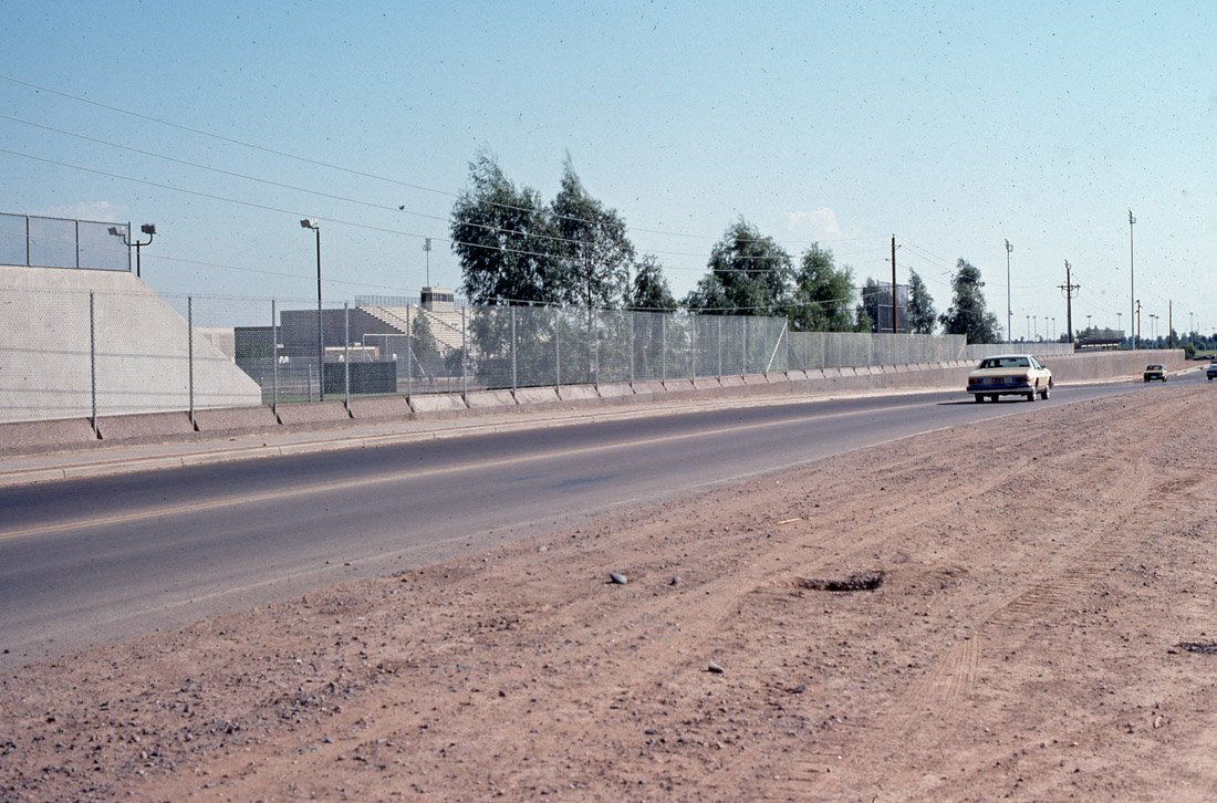 A wide, open road with a few cars driving away, a dirt shoulder on the side, and a chain-link fence running parallel to the road backed by trees and utility poles.