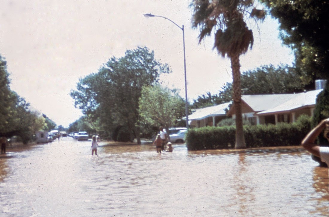 A flooded street with water covering the road, people walking through the flood, trees, parked cars, and a house in the background.