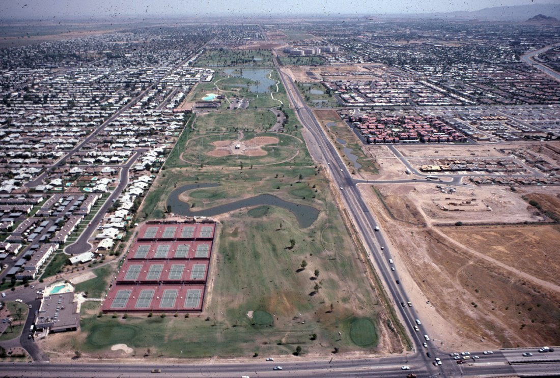 Aerial view of a city with a large park featuring a baseball field, tennis courts, a pond, and walking paths, surrounded by residential neighborhoods and highways.