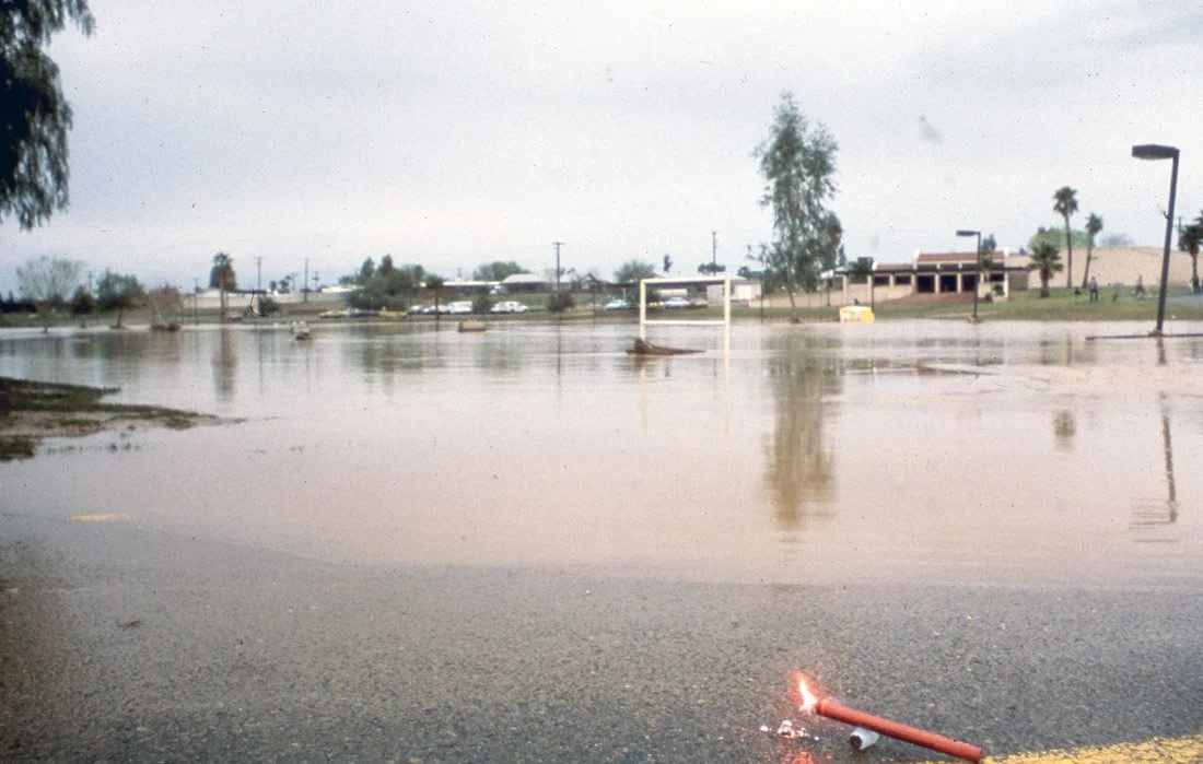 Flooded parking lot with water covering the ground, a soccer goal, and some trees and buildings in the background.