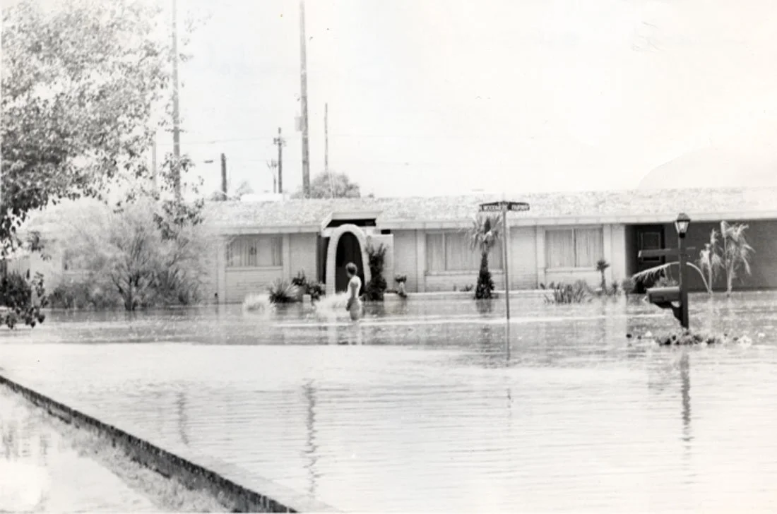 Suburban house surrounded by floodwaters, with a person standing in the water near the house and flooded street.