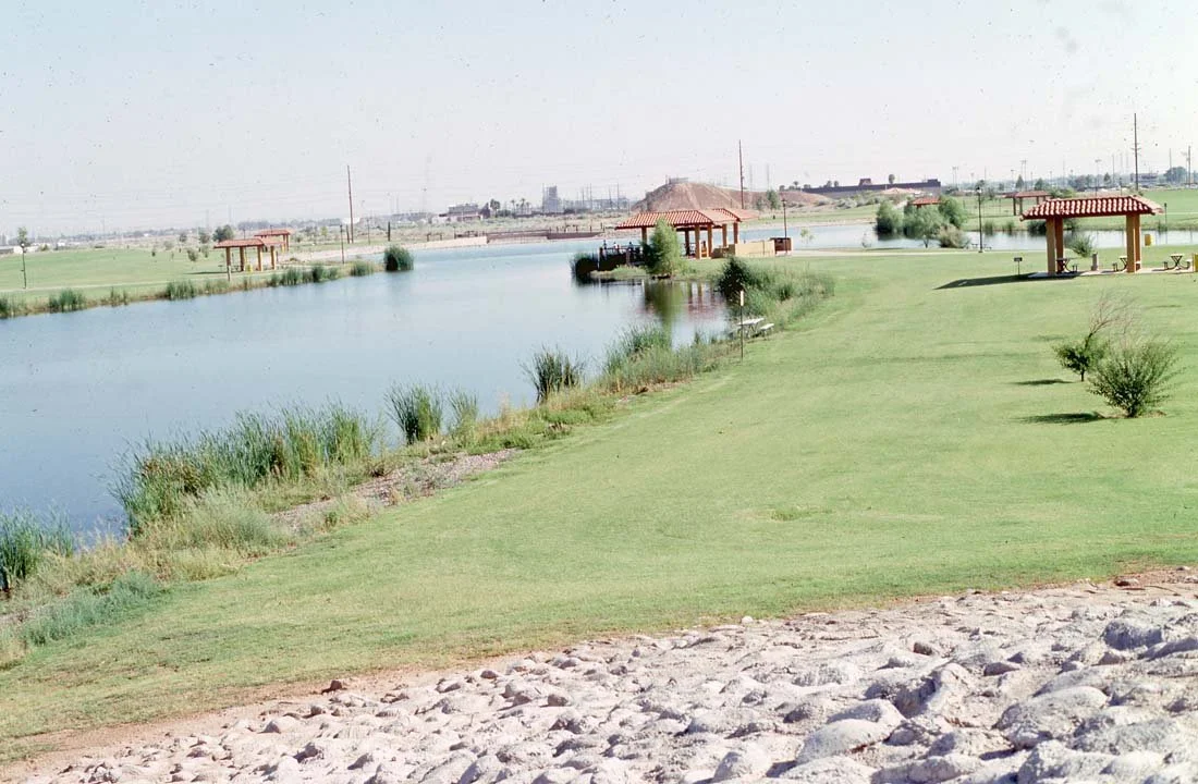 A park with a lake, grassy areas, small bushes, and a few pavilions with red roofs and picnic tables, during daytime.