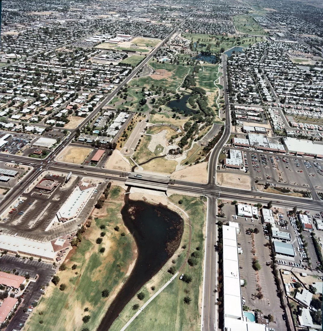 Aerial view of a city park with water features, surrounded by residential neighborhoods and commercial areas, including parking lots and roads.