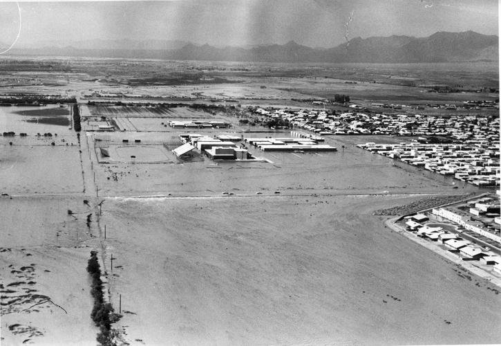 Aerial view of a flooded urban area with buildings and surrounding fields partially submerged. The landscape extends to mountains in the background under a cloudy sky.