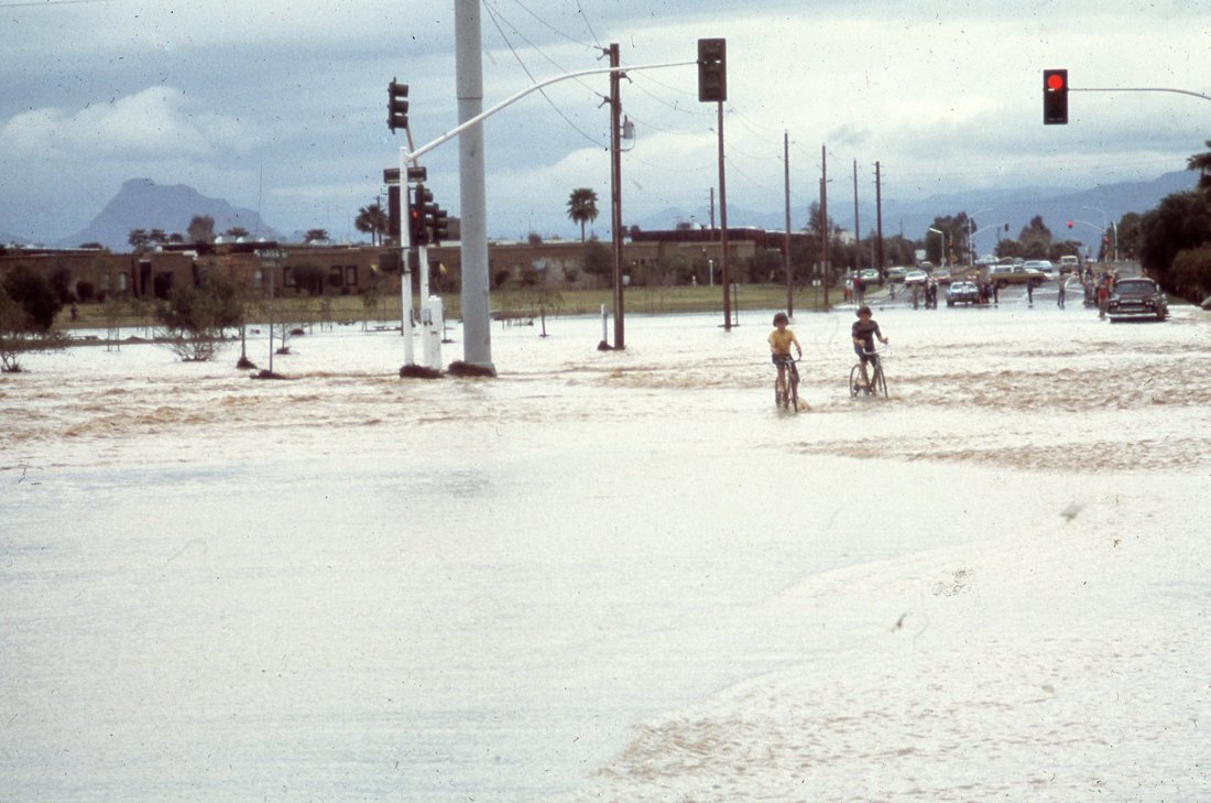 Two children riding bicycles across a street flooded with water after a flood, with traffic lights and vehicles in the background.