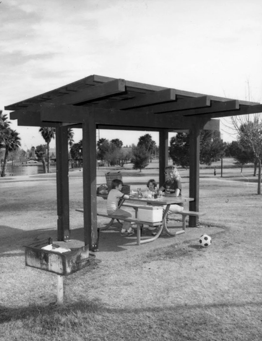 Three children and one adult sitting at a picnic table under a wooden shelter in a park, with a soccer ball on the ground nearby and trees in the background.