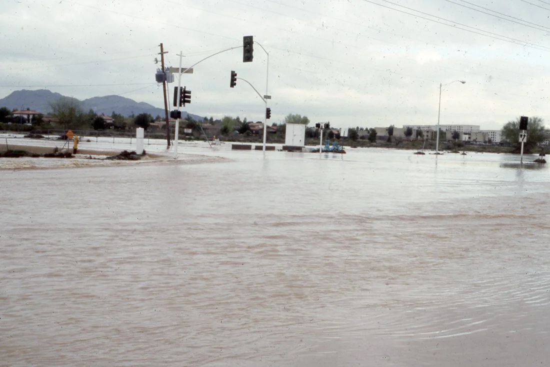 Flooded street with traffic lights and a utility pole, water covering the road, cloudy sky, mountains in the background.