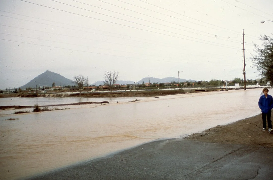 Flooded road with muddy water, a person in a blue jacket standing on the sidewalk, power lines, sparse trees, and distant mountains under an overcast sky.