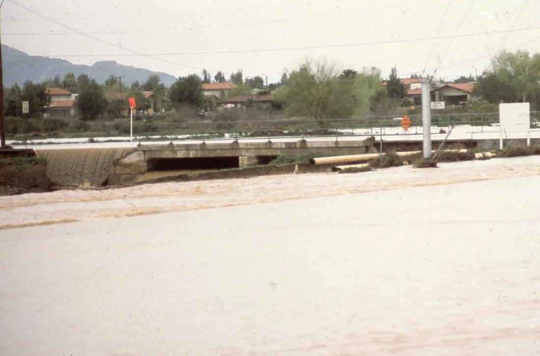 A flooded road with water covering the surface, a bridge or culvert underneath, and houses with trees in the background, indicating heavy rain or flooding.