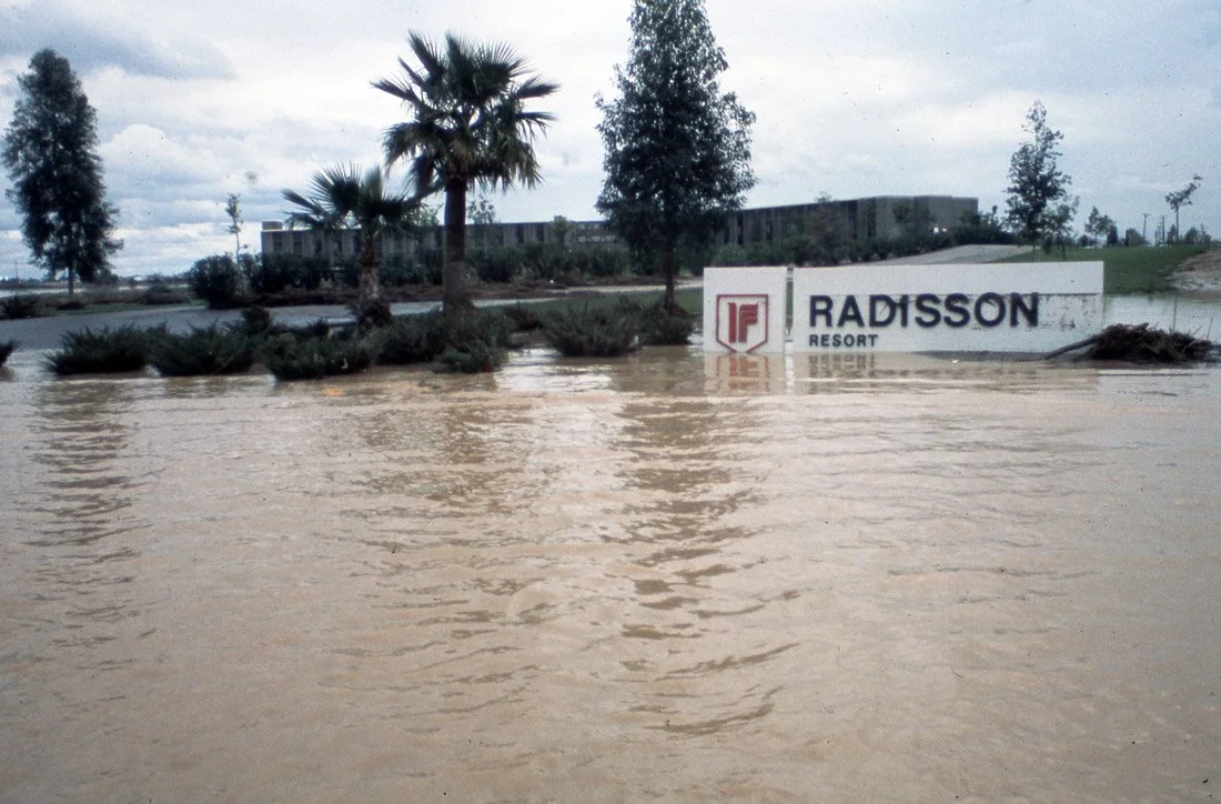 Flooded parking lot with water covering the ground and partially submerging a sign that reads 'Radisson Resort' with trees and clouds in the background.