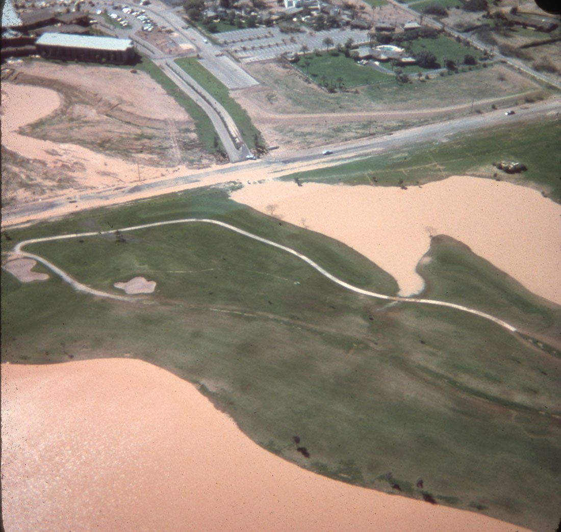 Aerial view of a park with walking paths, grassy areas, water bodies, and nearby roads and parking lots.