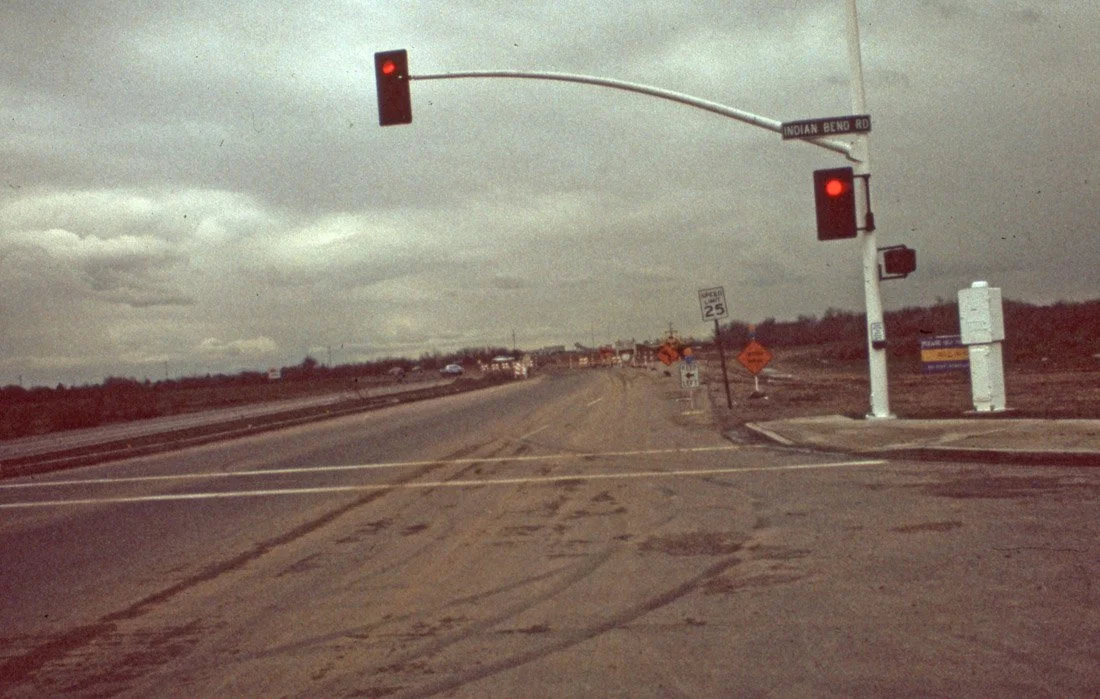 An intersection with traffic lights showing red signals, street sign reading Indian Bend Road, and roadwork signs indicating construction and reduced speed zone.