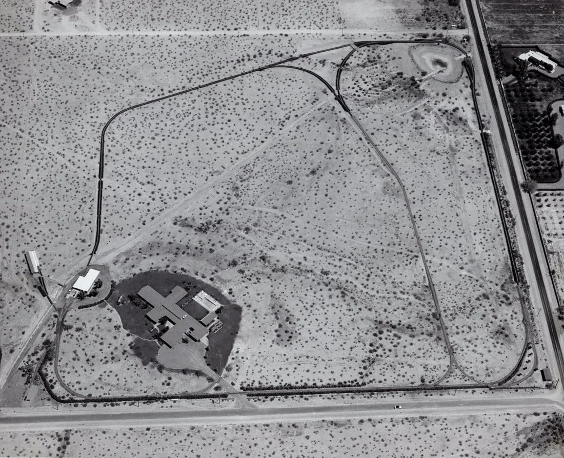 Aerial view of a large rectangular fenced area with a cross-shaped building in the center surrounded by grassy terrain and scattered trees. There are multiple roads and paths around and within the area.