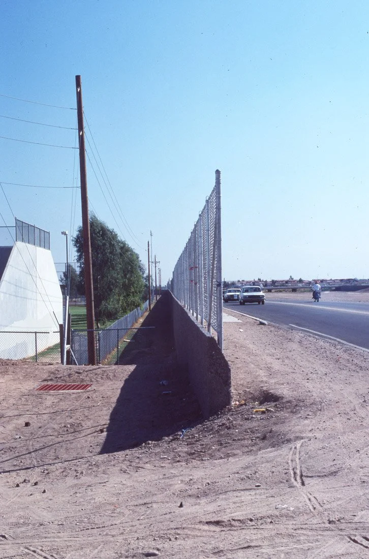 A stretch of road with a chain-link fence on the right side, and utility poles and trees on the left side. The road has cars and a cyclist, and the sky is clear and blue.