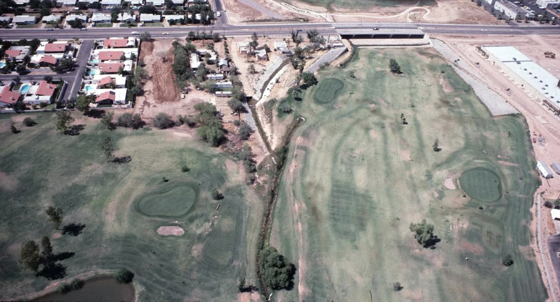 Aerial view of a golf course with sand traps and small ponds, next to a residential area and a highway.