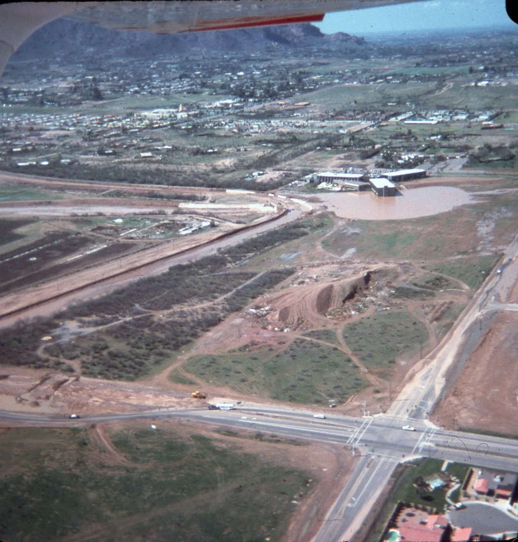 Aerial view of a developing area with construction sites, roads, and patches of land, overlooking a distant cityscape under a clear sky.