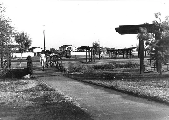Black-and-white photo of a park with a walking path, small bridges, trees, and people, some walking and some sitting, in a residential neighborhood with houses in the background.