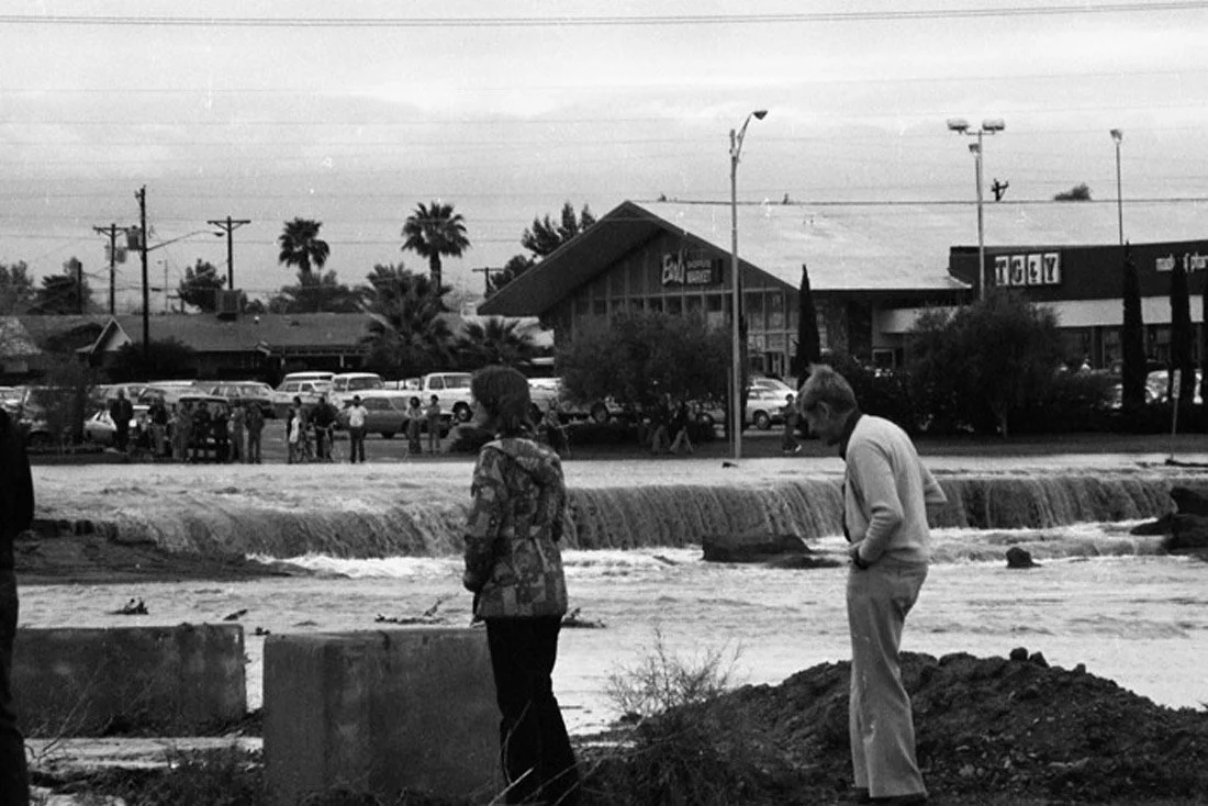People standing near a water feature with a small waterfall, surrounded by trees, buildings, and parked cars in the background, in a black and white photo.