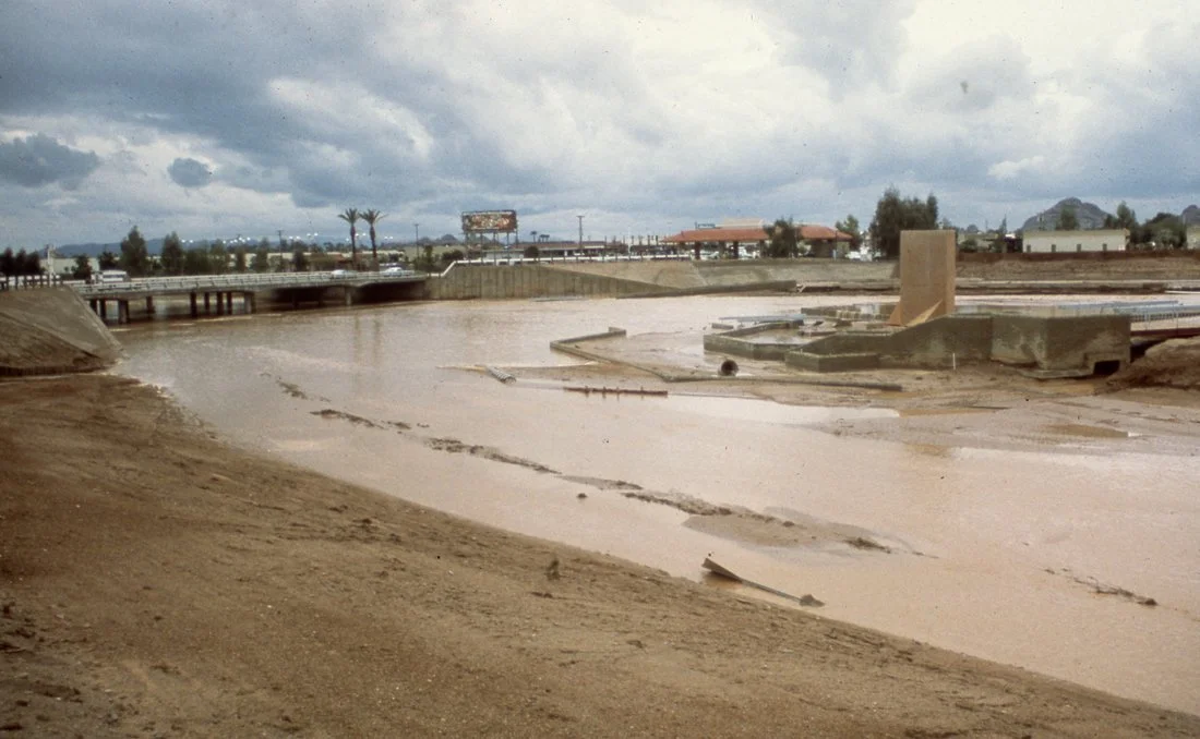 View of a flood-damaged area with muddy water, damaged structures, and a cloudy sky.
