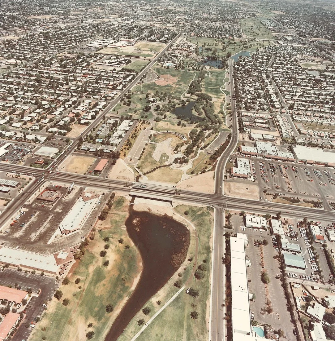 An aerial view of a park with water bodies, surrounding residential and commercial areas, highways, and parking lots.