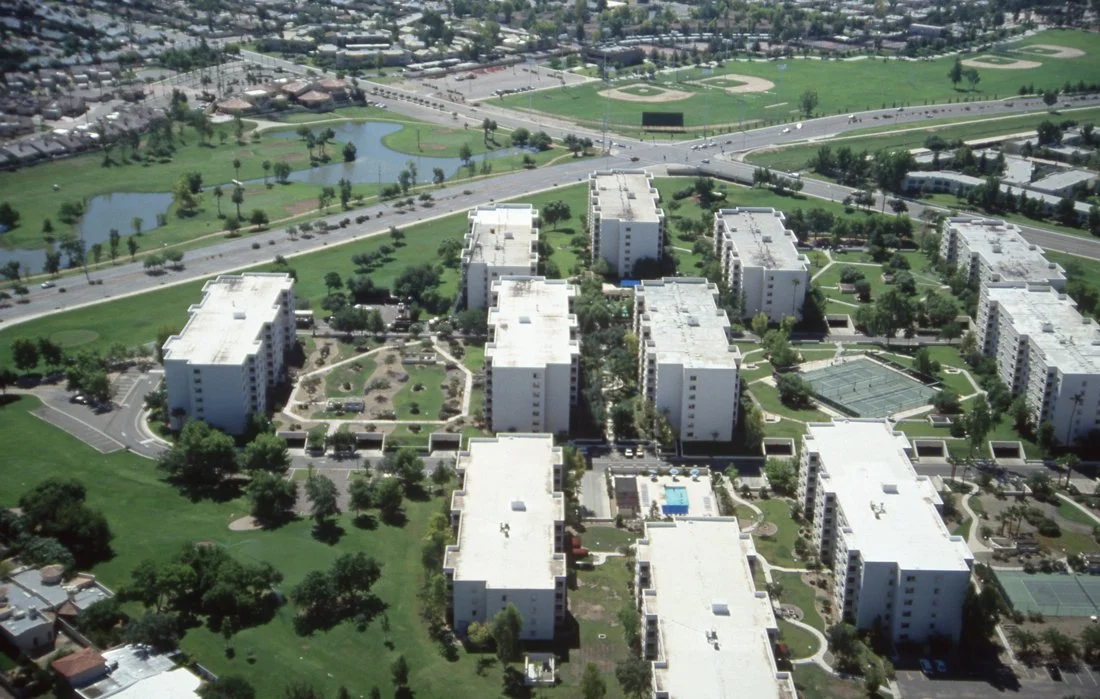 Aerial view of a residential apartment complex with multiple white buildings, landscaped greenery, a swimming pool, tennis courts, and nearby lakes and sports fields.