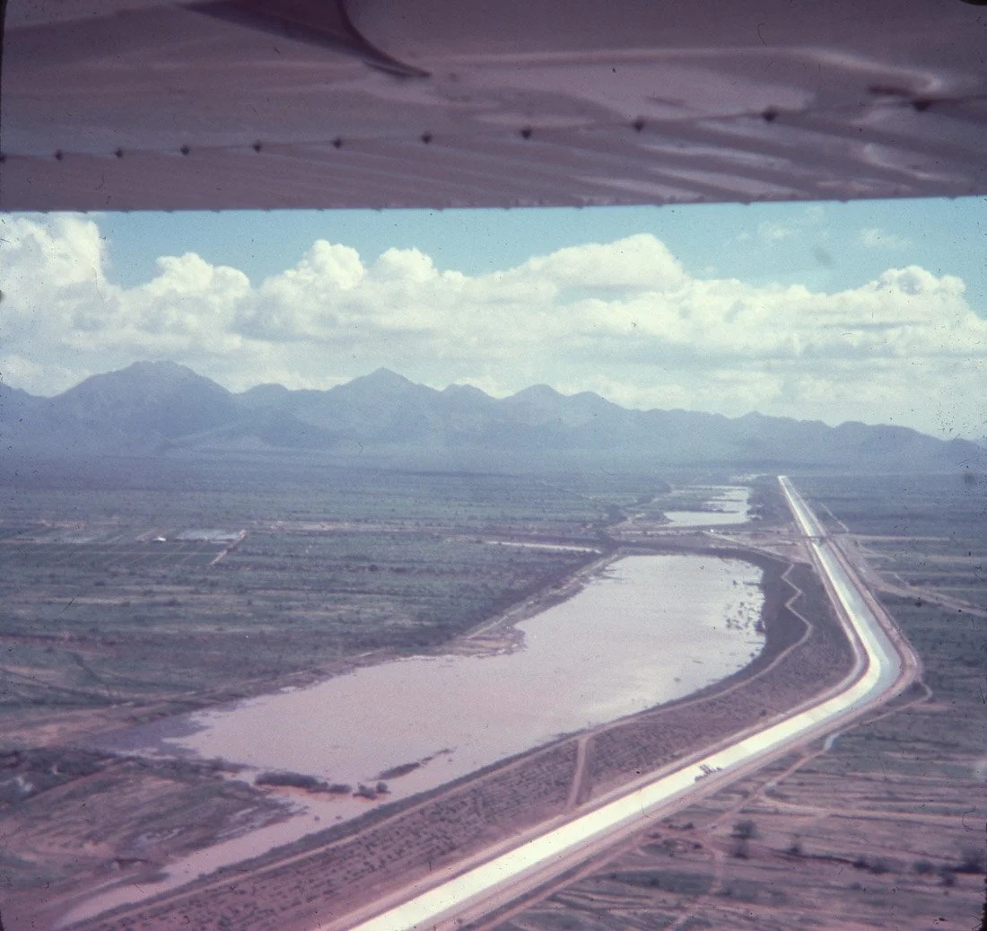 Aerial view from an airplane showing a landscape with mountains, fields, a body of water, and a long straight road or canal.