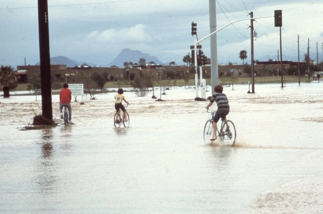 Three children riding bikes through a flooded street with traffic lights, utility poles, and mountains in the background.