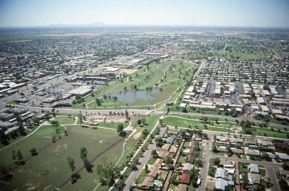 Aerial view of a city park with a pond, walking paths, green lawns, and surrounding residential and commercial areas.