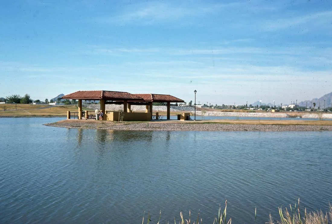 A small pavilion with a red roof built on a narrow strip of land surrounded by water, with a park and distant mountains in the background.