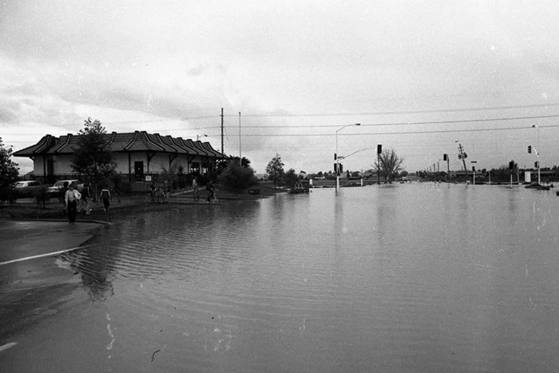Flooded street with water reaching the building's entrance, several people walking on a sidewalk, trees and power lines visible under a cloudy sky.