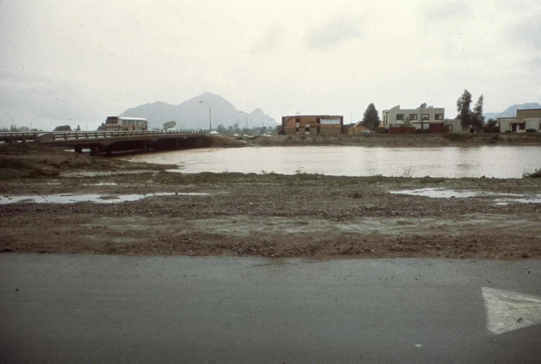 View of a bridge over a water body with houses and mountains in the background, under cloudy sky.