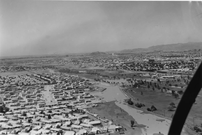 Aerial black-and-white photograph of a cityscape with a residential area on the left, a river running through the center, and open green spaces on the right, with mountains in the distance.