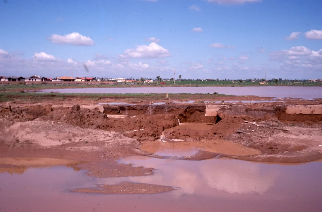 A muddy construction site near a body of water with houses and buildings in the background under a partly cloudy sky.