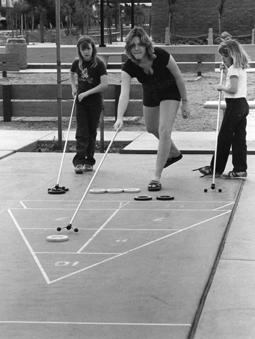 A woman and three young girls playing shuffleboard on an outdoor court, with the woman actively pushing a puck and the girls observing or preparing to play.