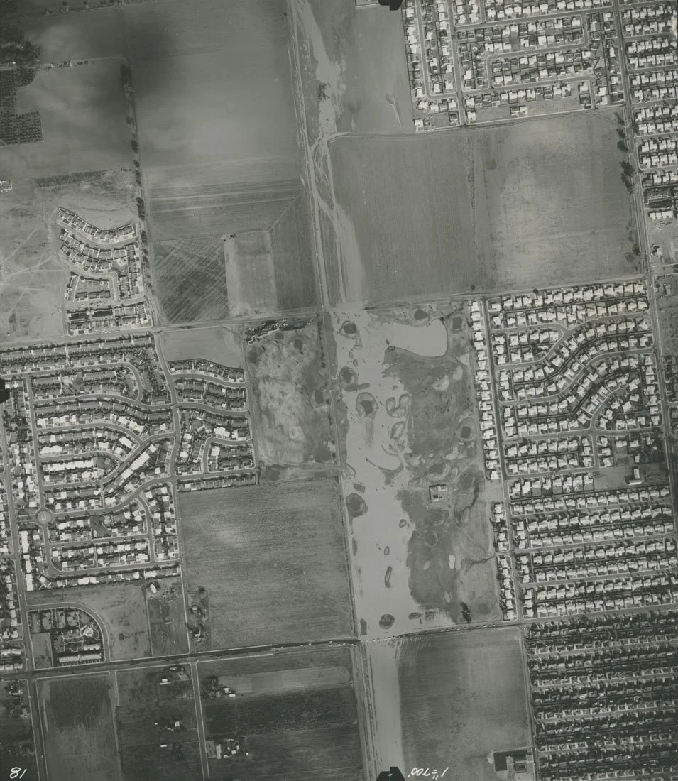 Aerial view of residential neighborhoods, agricultural fields, and open land, with a pond and winding roads in a suburban area.
