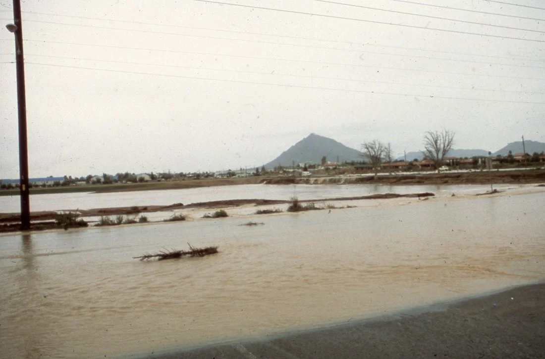 Flooded area with water covering the ground, debris floating, power lines, trees, and mountain in the background.