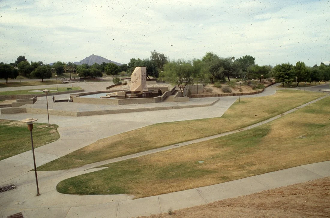 An empty park with walking paths, grassy areas, trees, benches, street lamps, and a monument in the background under a cloudy sky.