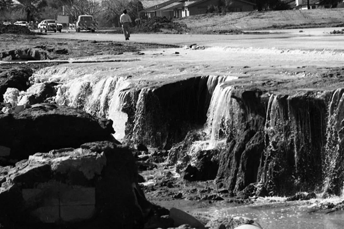 Black and white image of a flooded street with a fallen tree and a man walking in the background.