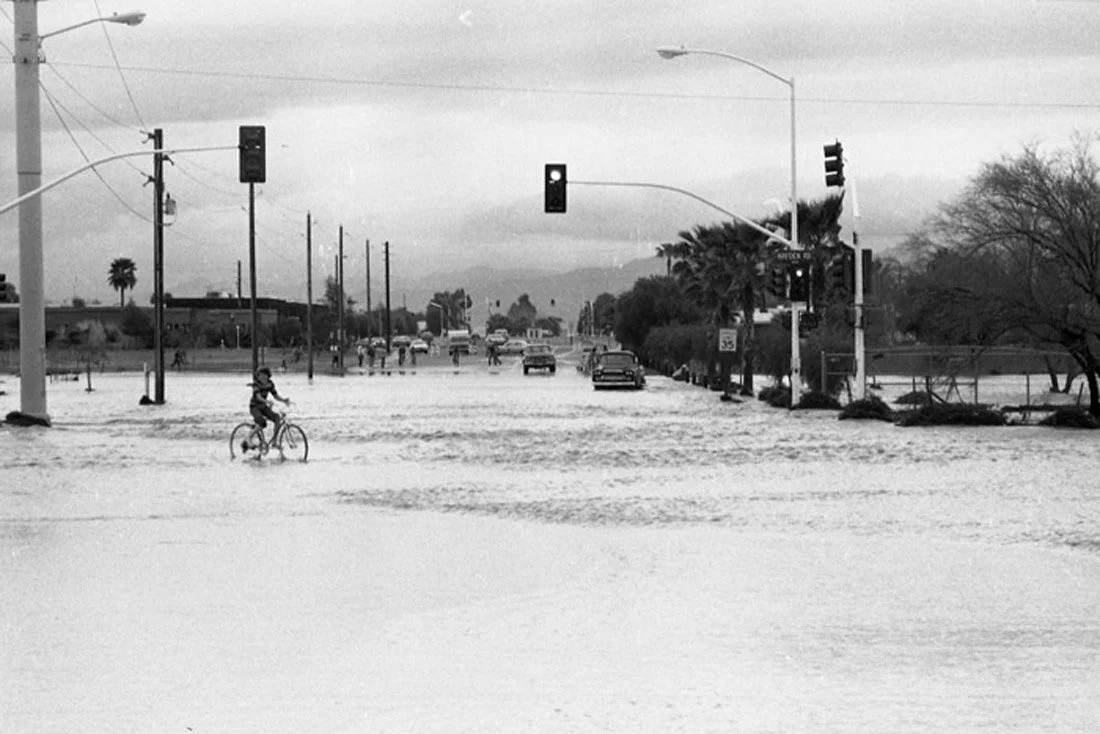 A flooded city street with cars and a child riding a bicycle through the water.