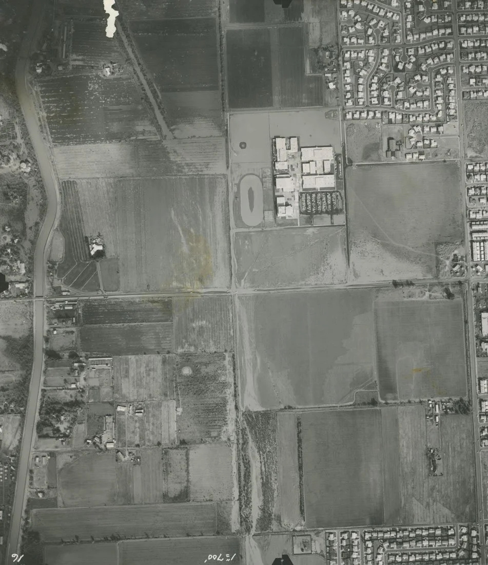 An aerial grayscale view of rural farmland, residential neighborhoods, and an educational or institutional complex with an athletic track, surrounded by roads and fields.