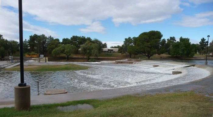 A park with a small man-made waterfall and flowing water, surrounded by grass, trees, and pathway, under a partly cloudy sky.