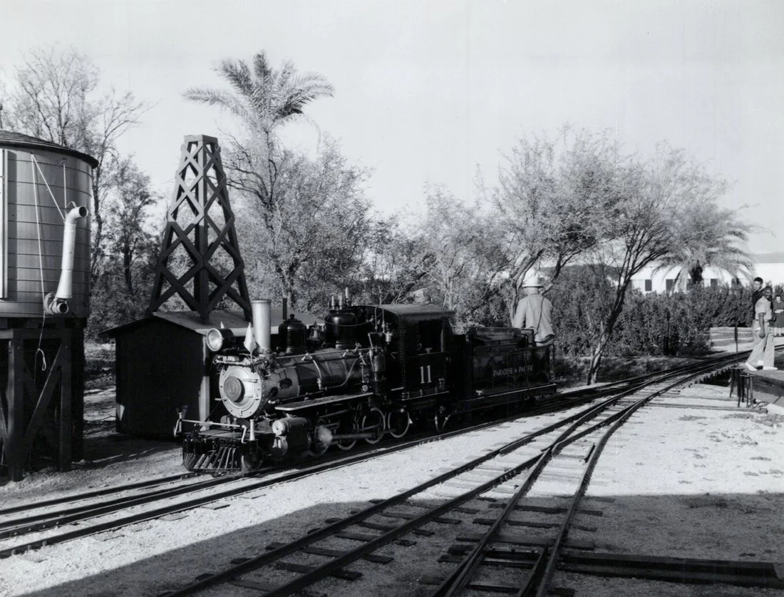 A small vintage steam train on track with trees and two people nearby.