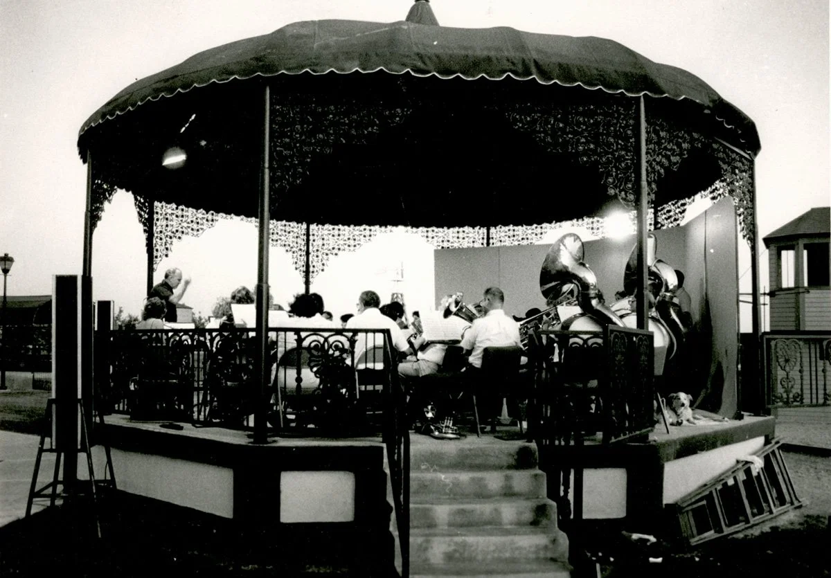 A black-and-white photo of a small outdoor bandstand with a musical ensemble playing instruments, including tubas and guitars, under a large umbrella or canopy.