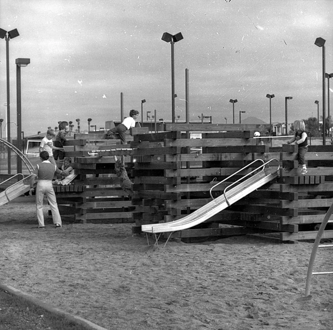 Children playing on wooden playground structure with slides and climbing areas, supervised by adult at a park with streetlights and overcast sky.